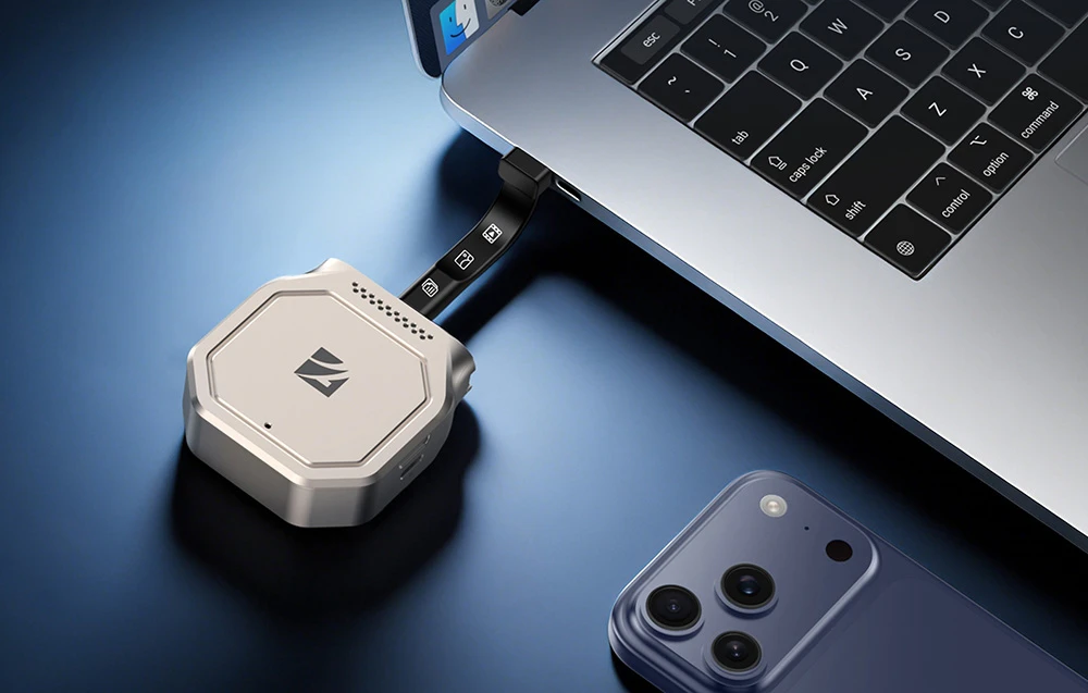 Un adaptor de hard disk Freewell argintiu conectat printr-un cablu USB-C flexibil la un laptop, lângă un smartphone așezat în apropiere