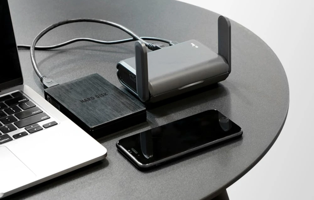 Router negru așezat pe o masă gri lângă un smartphone, un hard disk și un laptop