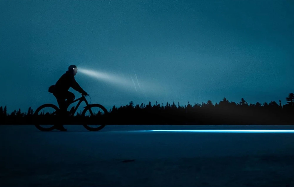 Silueta unui ciclist care pedalează noaptea. Lumina lanternei frontale îi luminează calea.