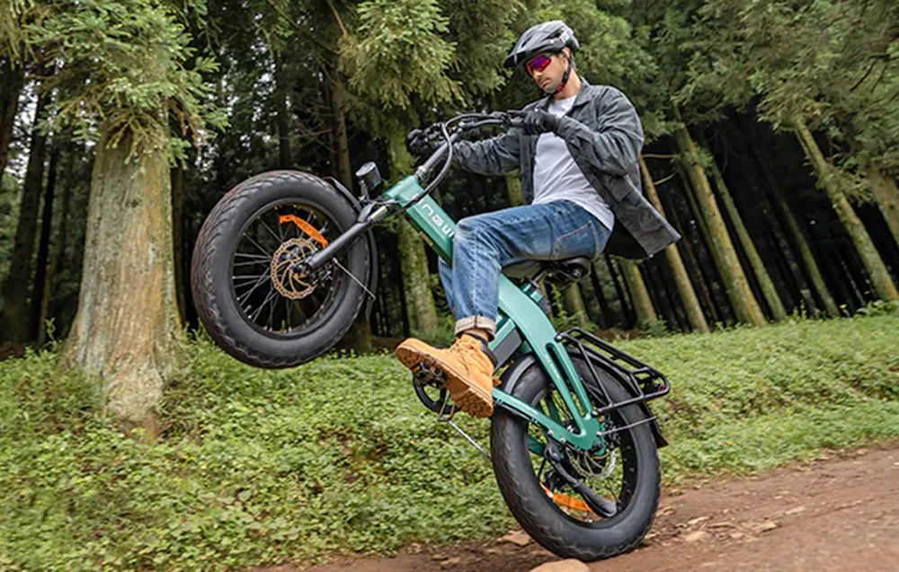 Un bărbat care poartă cască ridică roata din față a unei biciclete electrice în timp ce merge pe un drum forestier