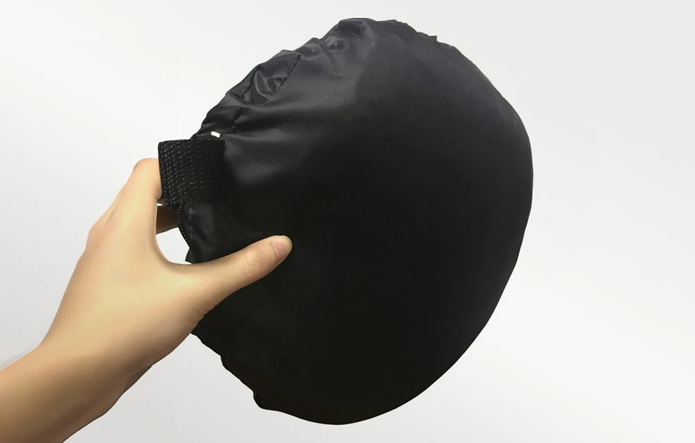 O mână ținând o husă rotundă, pliată, din țesătură neagră, pe un fundal de culoare deschisă