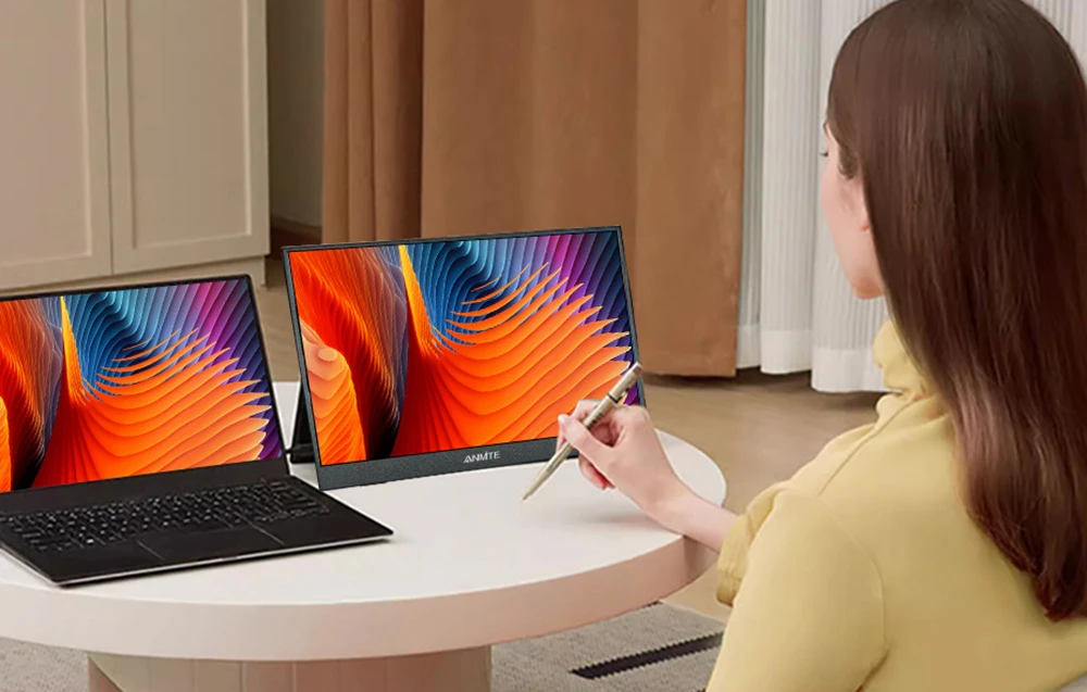 O femeie așezată la o masă rotundă folosește un stylus pe un ecran portabil amplasat lângă un laptop.