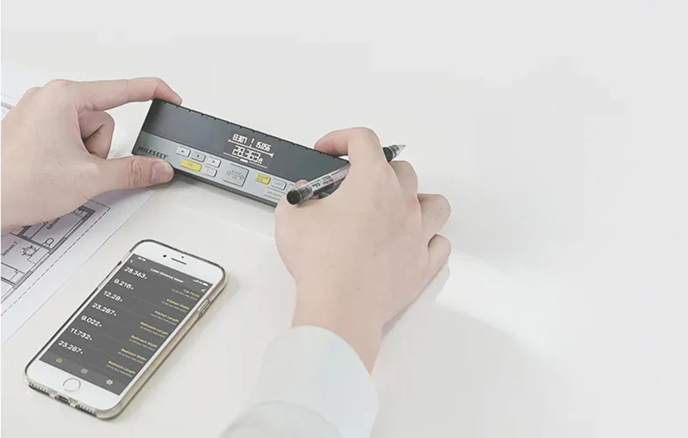 Telemetru laser ținut în mâini deasupra unui plan de construcție lângă un smartphone care afișează o listă de măsurători.
