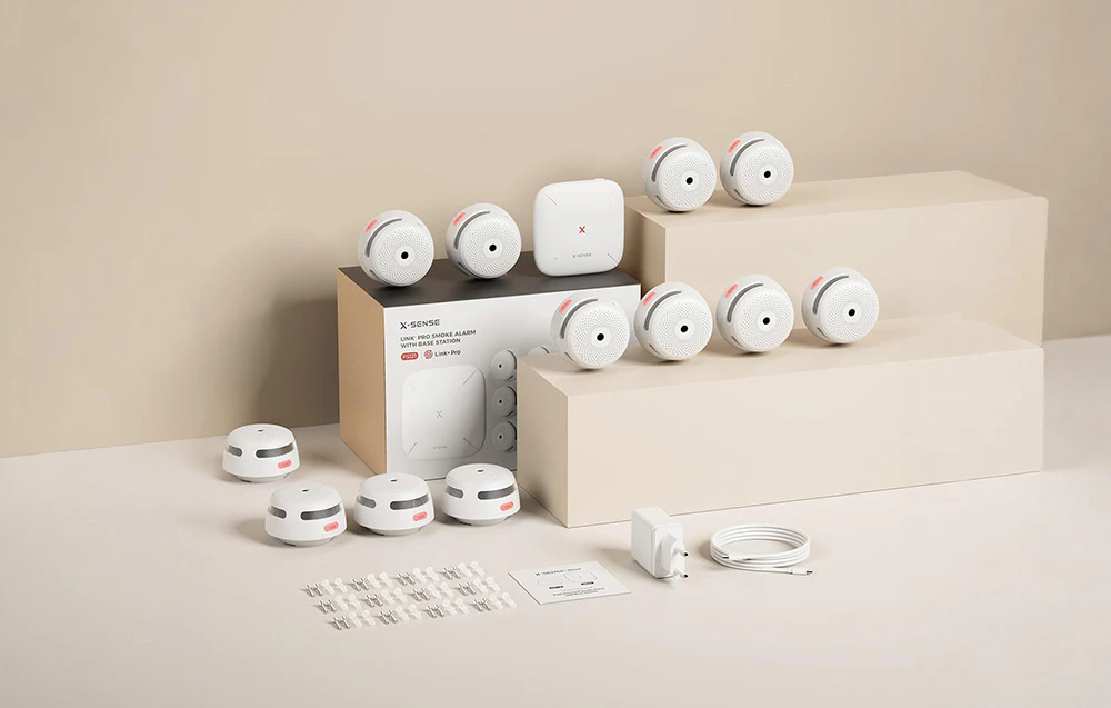 Set de produse X-Sense cu mai multe detectoare de fum, o stație de bază, o cutie și accesorii de montare pe un fundal bej