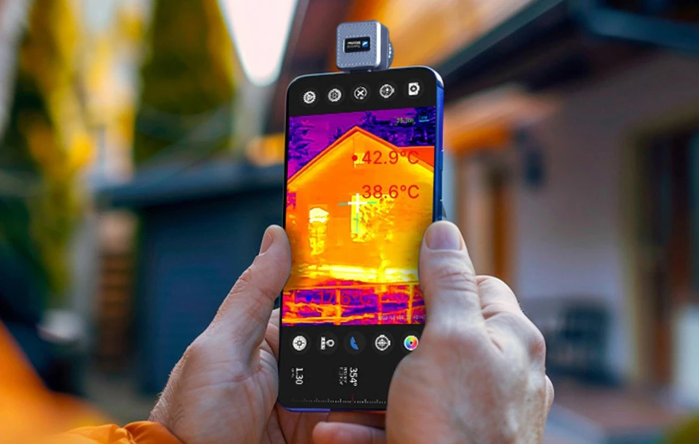 Mâinile țin un smartphone cu o cameră mică în partea de sus, care arată distribuția temperaturii pe peretele unei case.