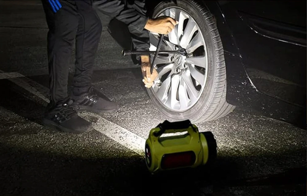 O lampă galbenă pe asfalt, lângă o persoană care folosește o cheie pentru a deșuruba o roată de mașină, iluminată de lumina albă a lămpii.
