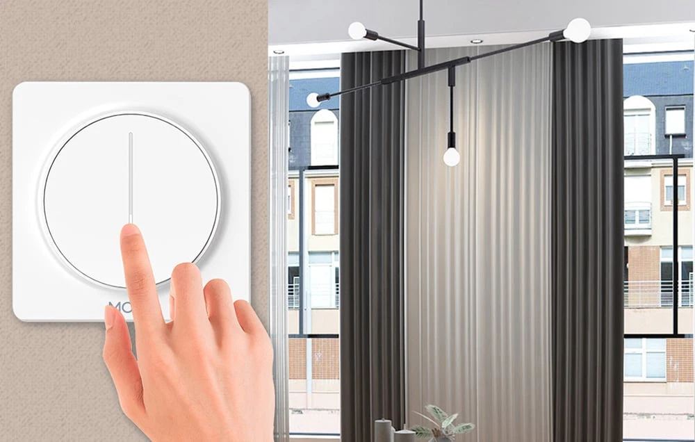 O mână atingând butonul rotund al dimmerului, pe fundalul unui candelabru modern într-o cameră luminoasă.