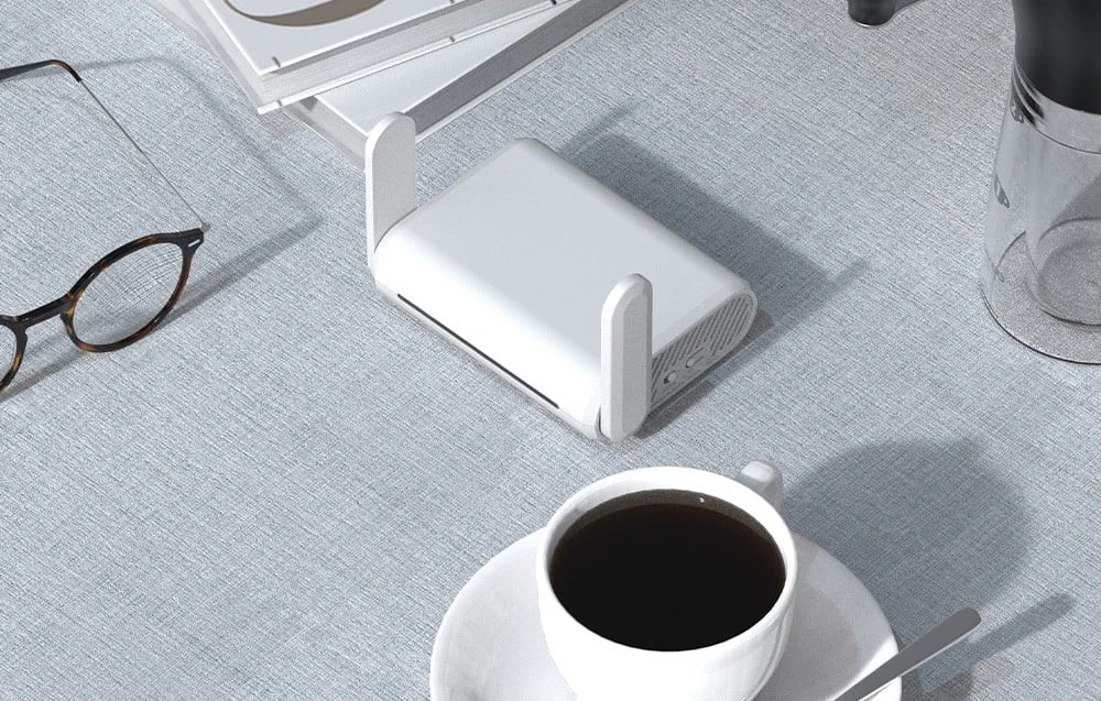 Un router alb cu antene așezat pe o masă gri, lângă o pereche de ochelari și o ceașcă de cafea