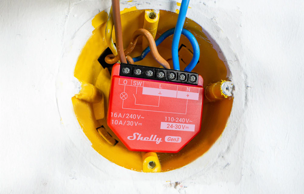 Un modul roșu Shelly 1PM Gen3 montat într-o cutie electrică în perete, cu firele conectate
