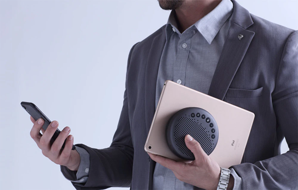 Un bărbat în costum ține un smartphone într-o mână și o tabletă sub braț, la care este atașat un difuzor de conferință rotund