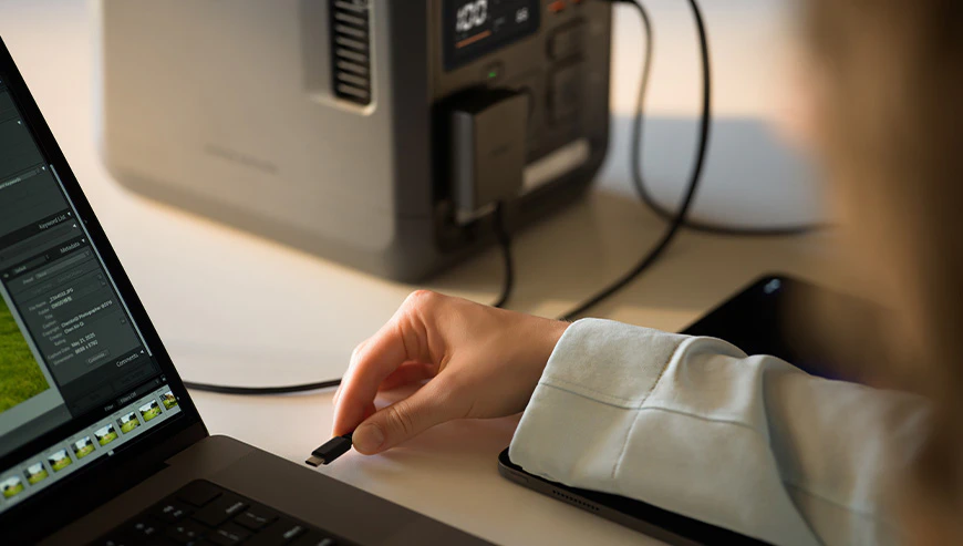 O persoană ține un cablu USB-C negru lângă un laptop cu un program de editare foto deschis, iar în fundal se vede stația de alimentare DJI Power 1000 Mini, conectată la o sursă de alimentare