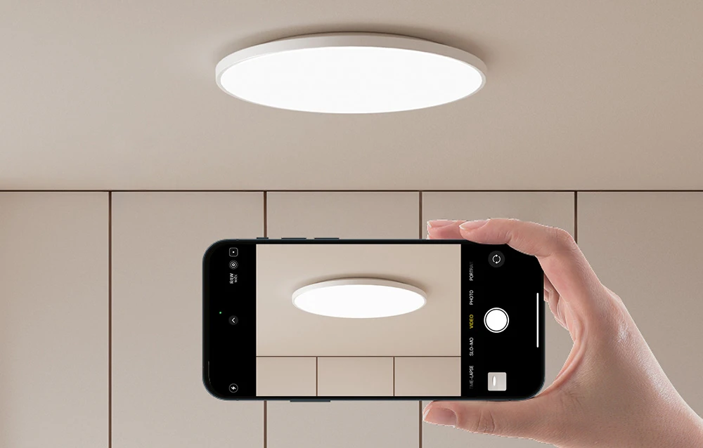 O mână care ține un smartphone cu camera îndreptată spre lampa de tavan Yeelight aprinsă.