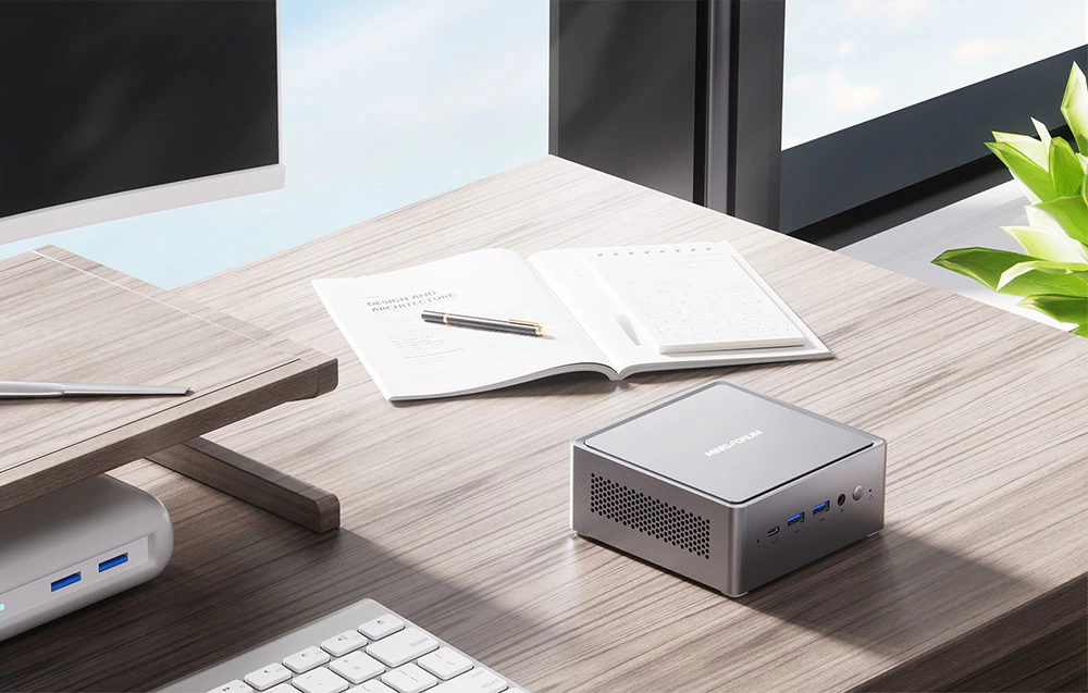 Birou într-o încăpere luminoasă cu mini PC, două notebook-uri deschise, tastatură și monitor în picioare lângă o fereastră cu o plantă