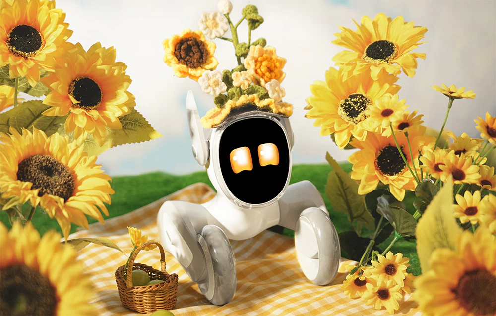 Robotul interactiv Loona într-un costum tricotat cu floarea-soarelui, așezat pe un covoraș galben de picnic, înconjurat de flori de floarea-soarelui