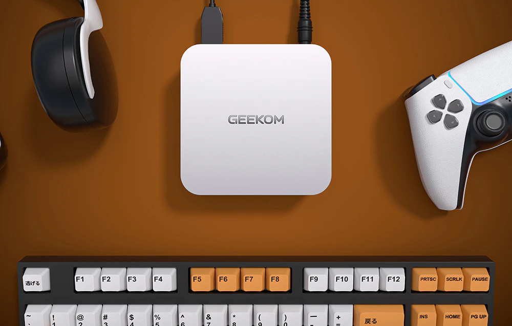 Mini PC Geekom pe un birou lângă un gamepad, tastatură și căști