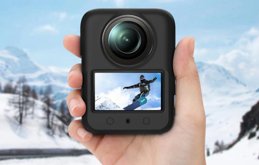Mâini care țin o cameră neagră de 360 de grade cu imaginea unui snowboarder pe ecran, pe un fundal montan