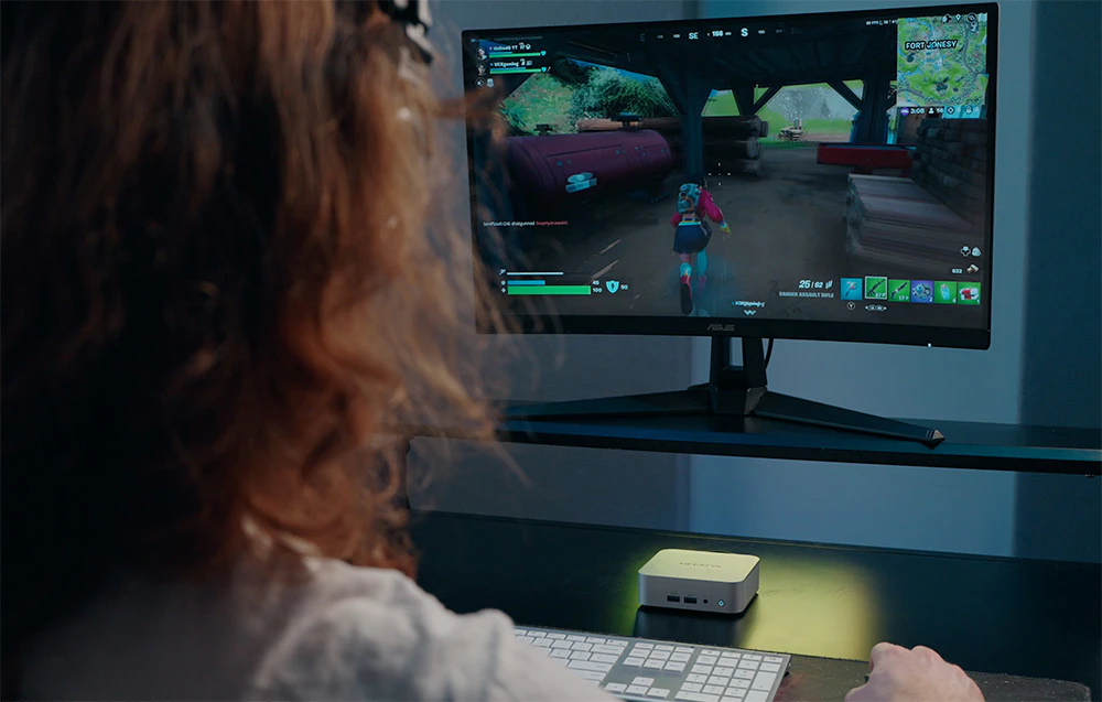 O persoană care joacă un joc pe computer pe un monitor mare, lângă care se află un mini PC cu iluminare din spate pe birou.