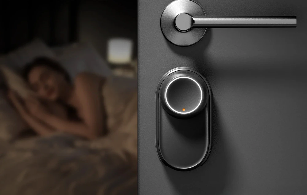 Încuietoare SwitchBot cu iluminare de fundal LED albă montată pe ușa întunecată a unui dormitor, cu o persoană dormind în fundal