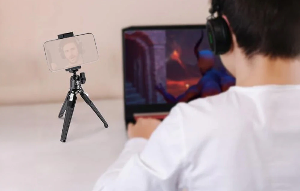 O persoană care poartă căști stă în fața unui laptop, lângă un mic trepied de birou cu un smartphone pe care se vede fața streamerului.