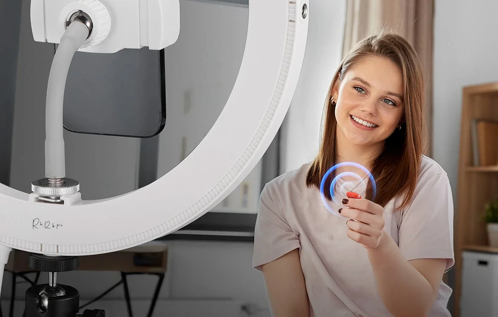 O fată zâmbitoare ține în mână o telecomandă Bluetooth fără fir pentru a declanșa camera de pe telefonul ei, stând lângă un inel luminos.