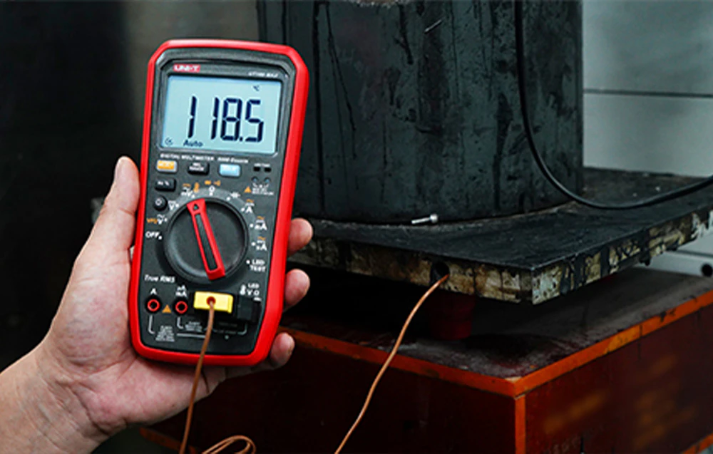 O mână în mănușă roșie ține un multimetru UNI-T care afișează 49,77 Hz, poziționat lângă un motor și un invertor.