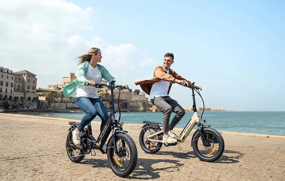 O femeie și un bărbat se deplasează cu biciclete electrice de-a lungul unei promenade de pe malul mării