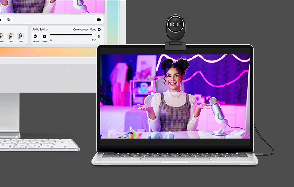 O cameră web atașată la un laptop care afișează o transmisiune live găzduită de o femeie zâmbitoare, cu un monitor și o tastatură lângă ea