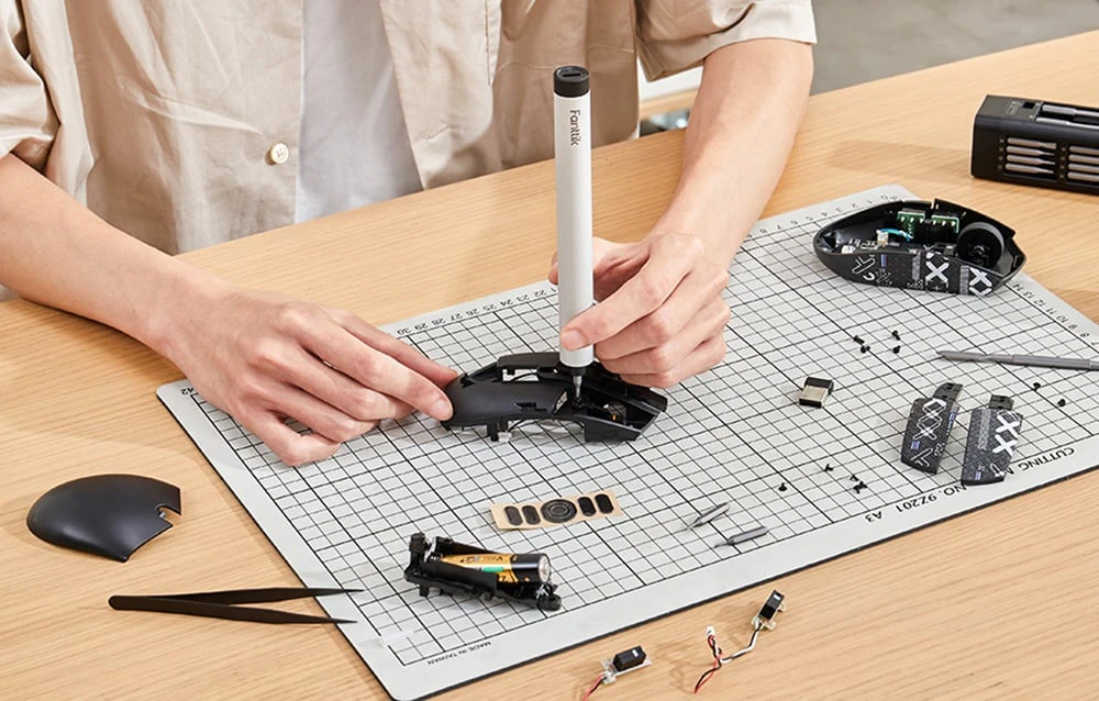 Un bărbat stă la biroul său și repară un mouse de calculator cu o șurubelniță de precizie.