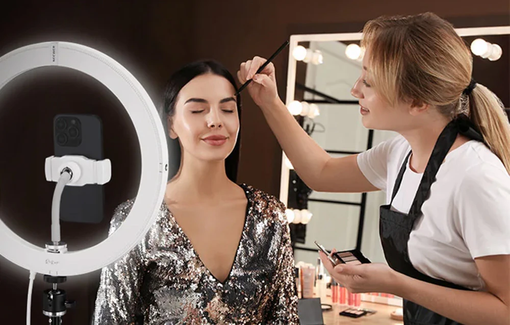O vizită la un artist de machiaj care aplică machiajul sprâncenelor unui model îmbrăcat într-o rochie strălucitoare, iluminat de un inel luminos alb Neewer.