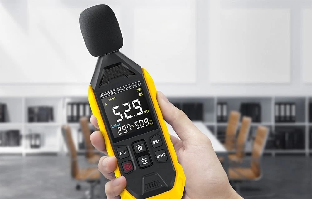O mână ține decibelmetrul cu apărătoare de microfon și afișaj care arată 52,9 dB, la birou.
