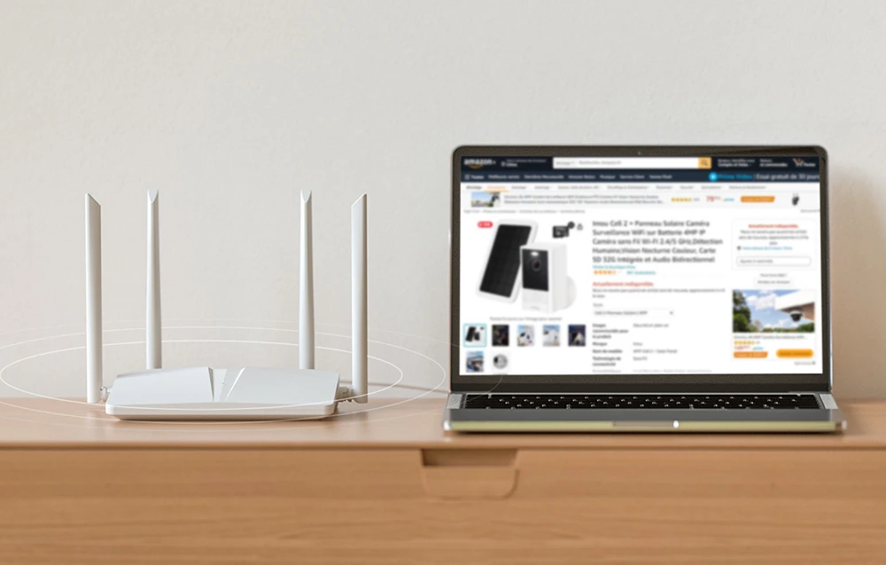 Un router alb cu patru antene așezat pe un dulap, lângă un laptop care afișează o pagină Amazon cu kitul de camere Imou Cell 2