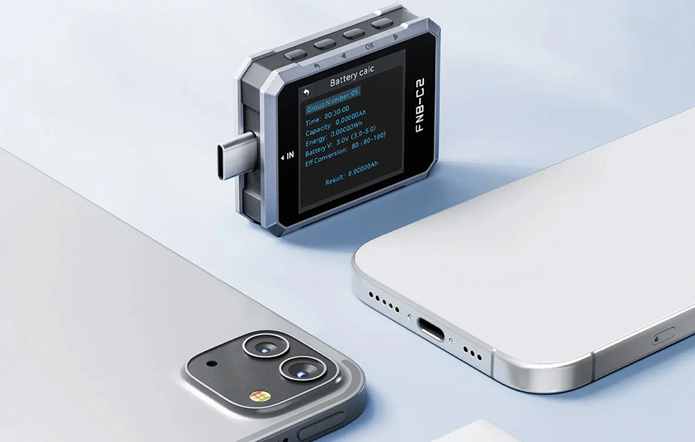 Un tester USB cu funcția de calcul al capacității bateriei afișată pe ecran, așezat lângă o husă de smartphone pe un fundal de culoare deschisă.