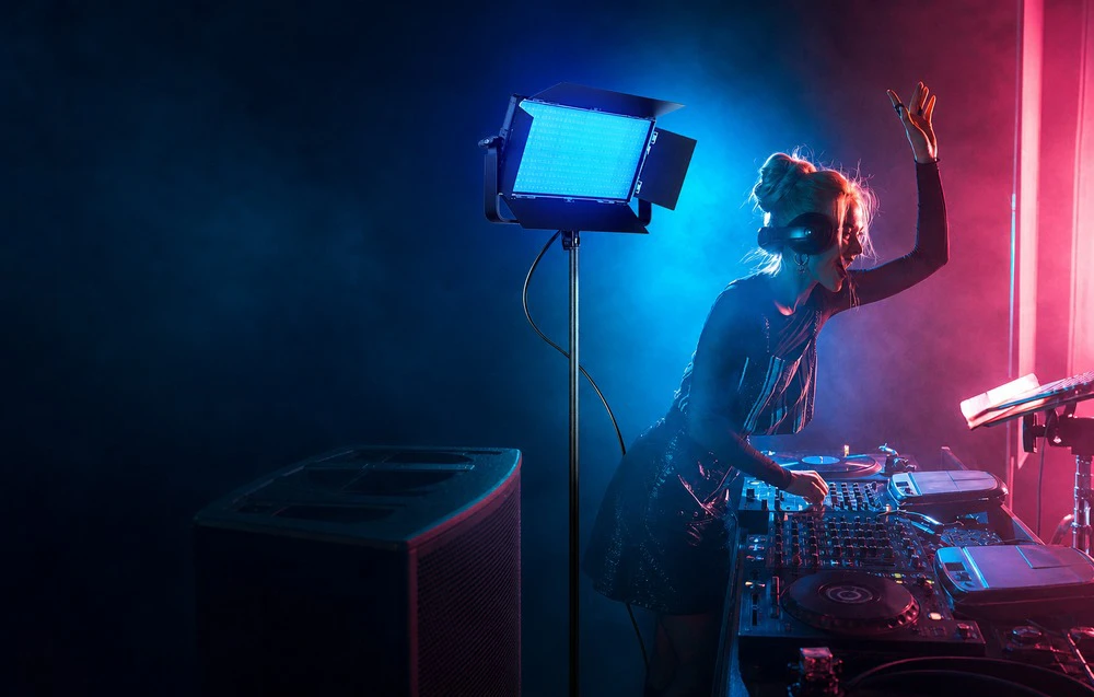 O DJ-iță mixează la o consolă de muzică într-un studio întunecat, iluminat de lumina albastră și roz a unei lămpi.
