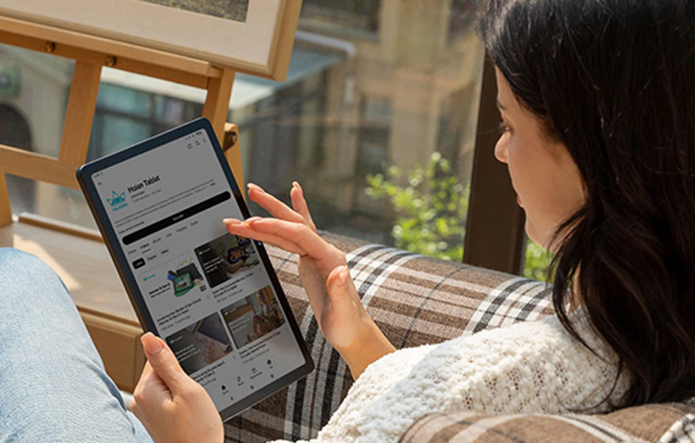 Femeie navigând pe site-ul Huion Tablet pe tabletă, stând confortabil lângă fereastră