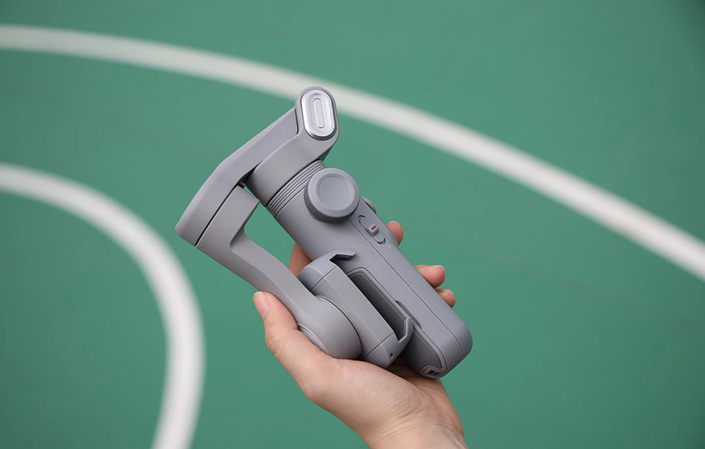 O mână ține un gimbal compact, pliat, pe fundalul unui teren de sport verde.