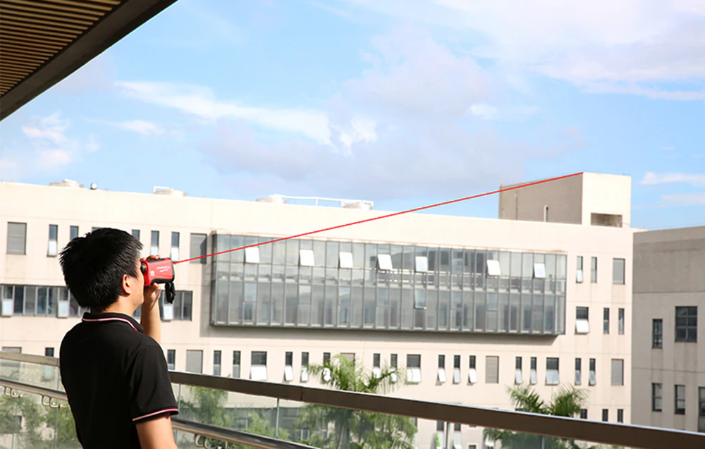 Un bărbat aflat pe un balcon folosește un telemetru, îndreptând o linie laser roșie către acoperișul unei clădiri îndepărtate.