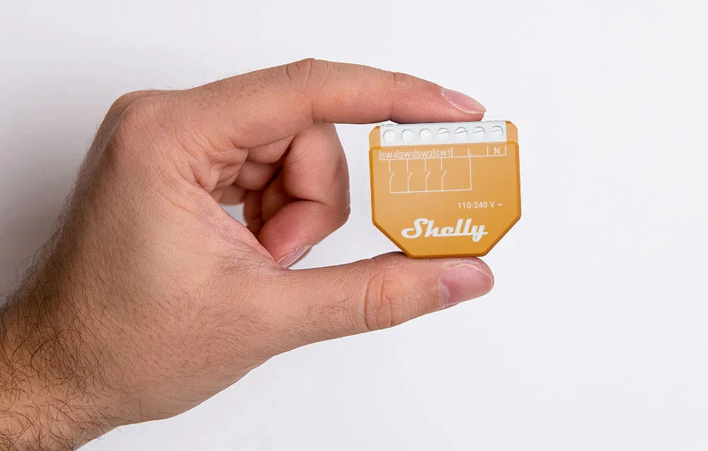 O mână care ține un modul Shelly portocaliu cu logo-ul Shelly și conectori pe un fundal alb