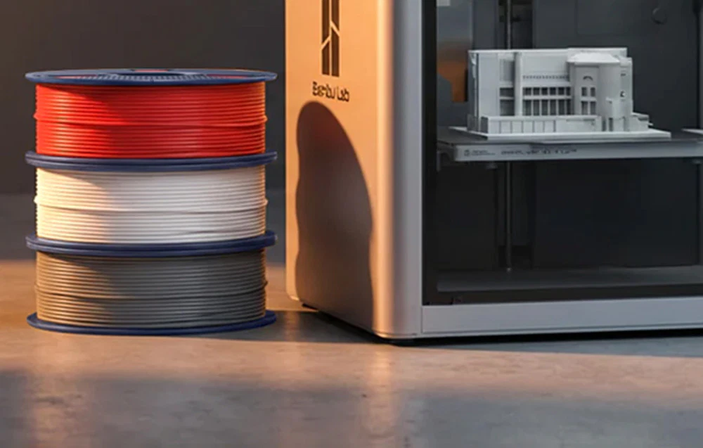 Trei bobine de filament colorat așezate lângă o carcasă metalică de culoare deschisă, în interiorul căreia se află un model de clădire imprimat.