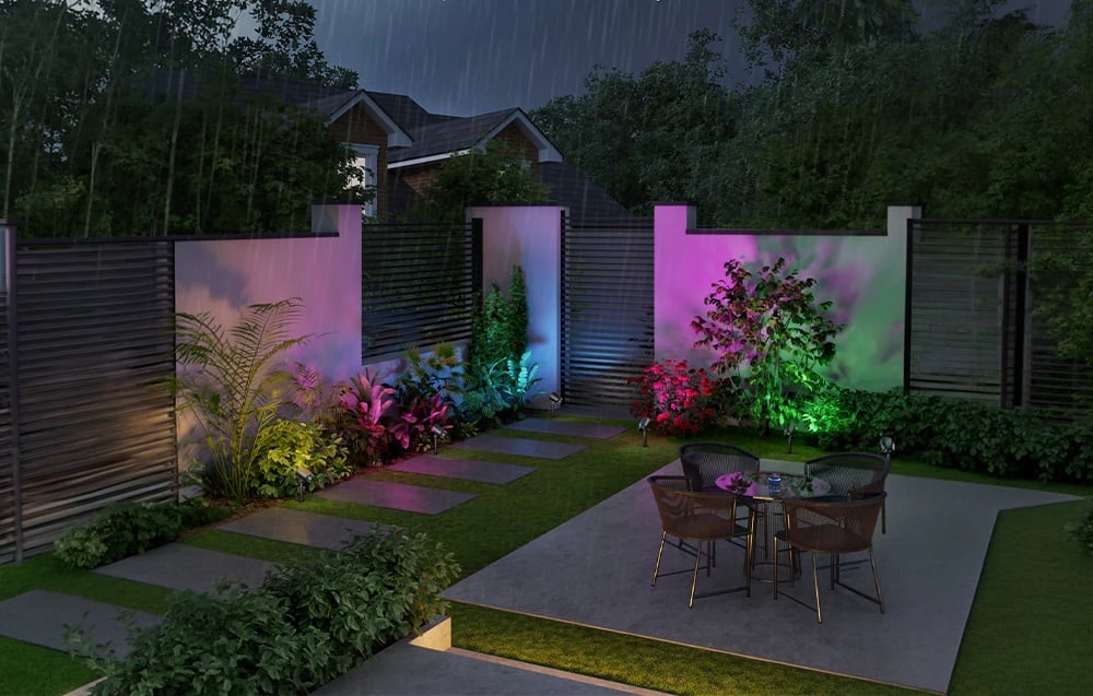 O grădină modernă cu mobilier de terasă sub ploaie, iluminată de lumina colorată a proiectoarelor de grădină.