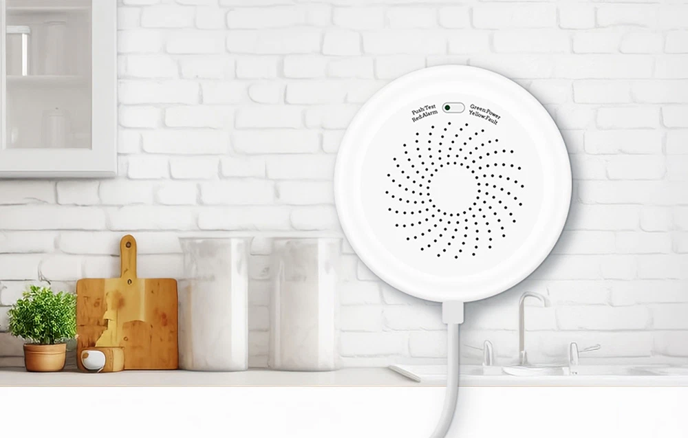 Detector de gaz alb, rotund, cu cablu de alimentare, pe fundalul unei bucătării luminoase cu un perete din cărămidă albă