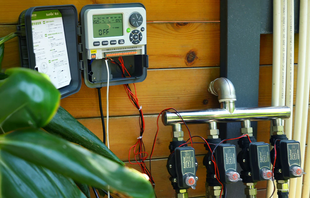 Un controler de irigare RainPoint® deschis, montat pe un perete din lemn, conectat prin cabluri la patru electrovalve instalate pe un colector metalic de apă.