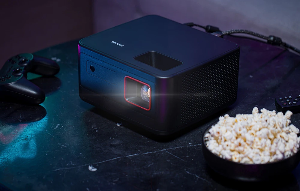 Un proiector PHILIPS negru cu lentilă iluminată din spate se află pe o masă întunecată lângă un bol de popcorn și un controler de jocuri