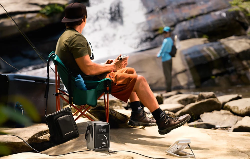 O persoană așezată pe un scaun de camping lângă o cascadă cu o stație electrică portabilă pe sol