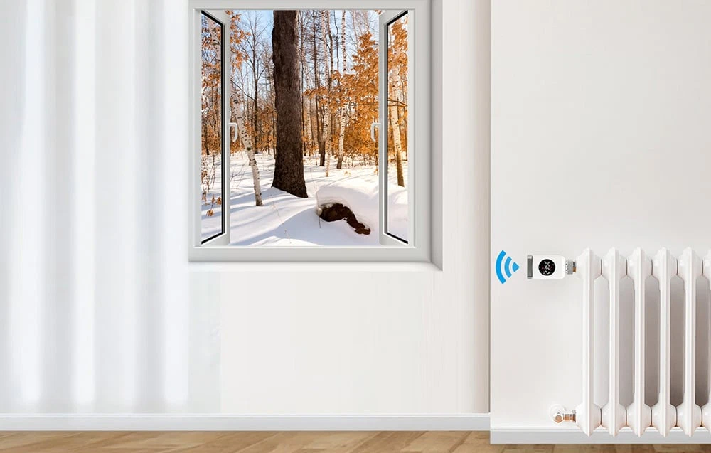 Un cap termostatic inteligent montat pe un radiator într-o cameră luminoasă, cu vedere la o pădure de iarnă în afara ferestrei.