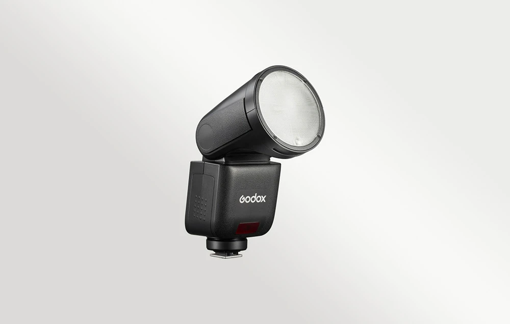 Un bliț Godox V1 S negru cu capul rotund orientat în sus și înclinat, pe un fundal de culoare deschisă