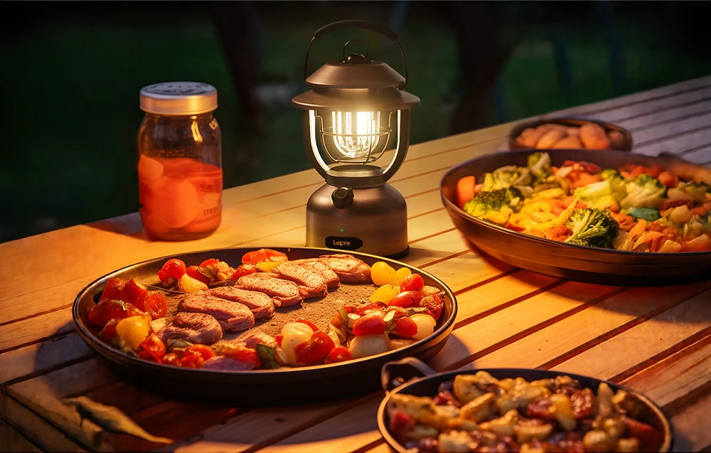 O lampă de camping aprinsă stă pe o masă de grădină din lemn, lângă farfurii cu mâncare