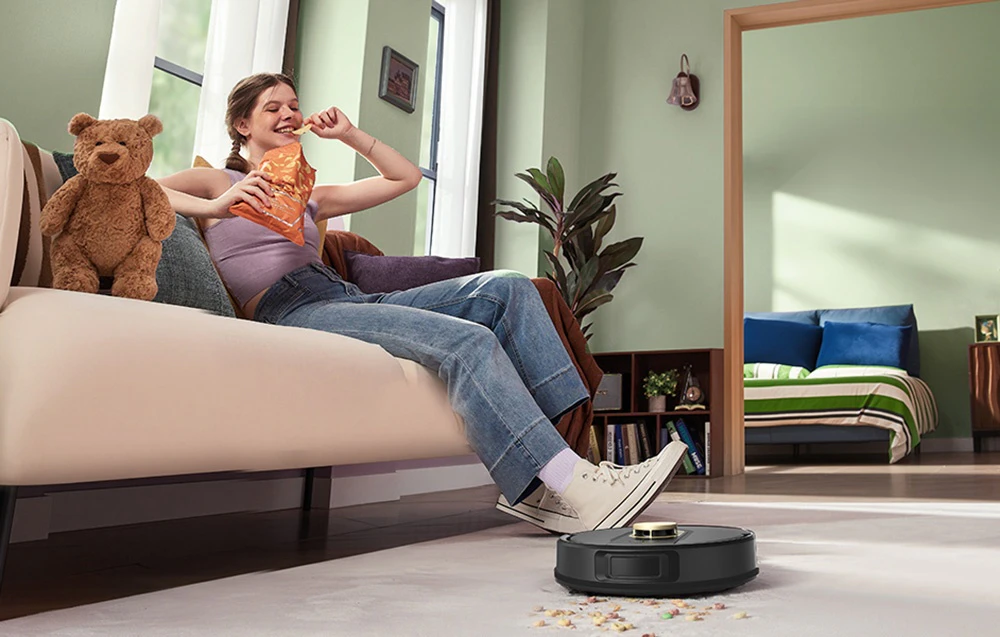 O fetiță care mănâncă chipsuri pe canapea și un robot aspirator negru care aspiră firimituri de pe covor