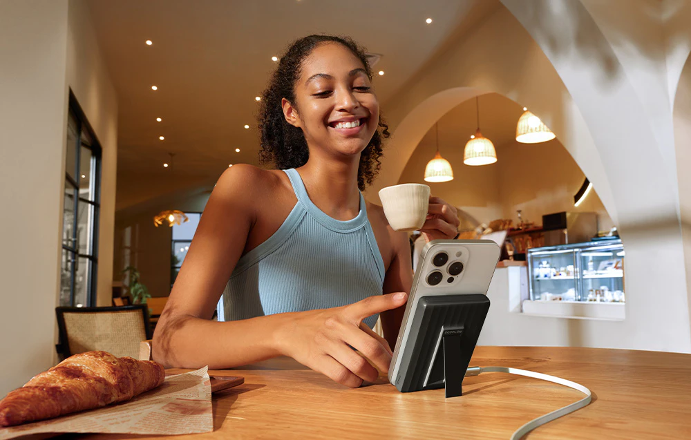 O femeie zâmbitoare într-o bluză albastră stă într-un restaurant și ține în mână o ceașcă. Pe masă se află un croissant și un telefon conectat la un powerbank magnetic cu suport.