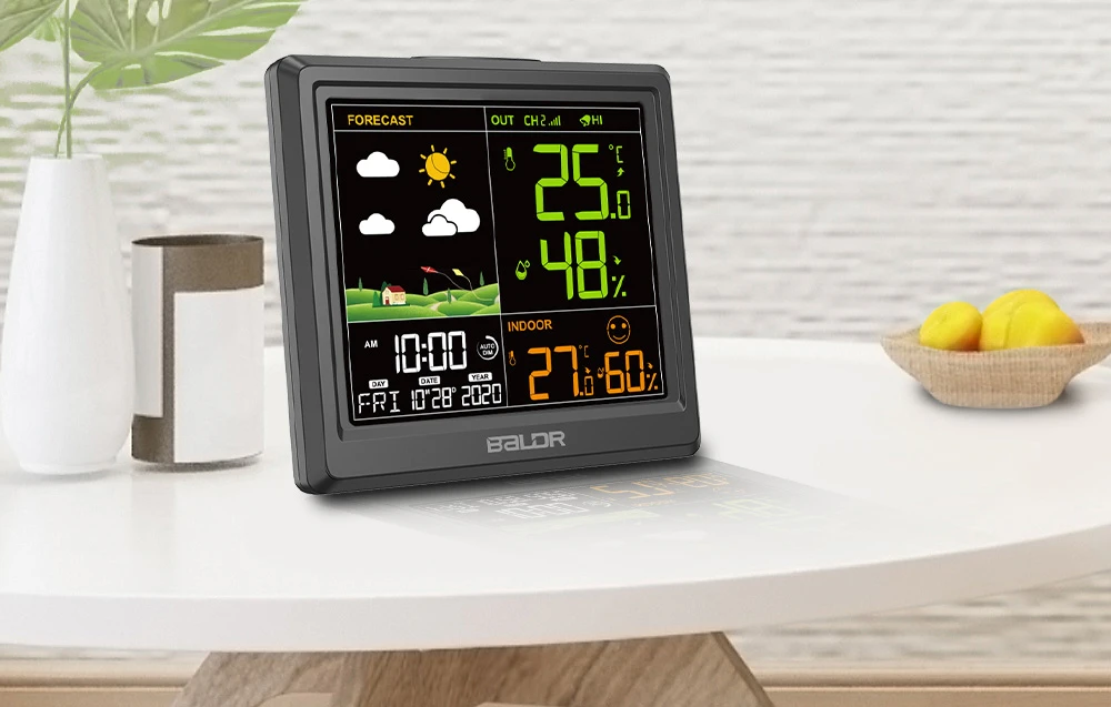 O stație meteo neagră așezată pe o masă de culoare deschisă, lângă o vază cu o plantă; în dreapta, un bol cu fructe