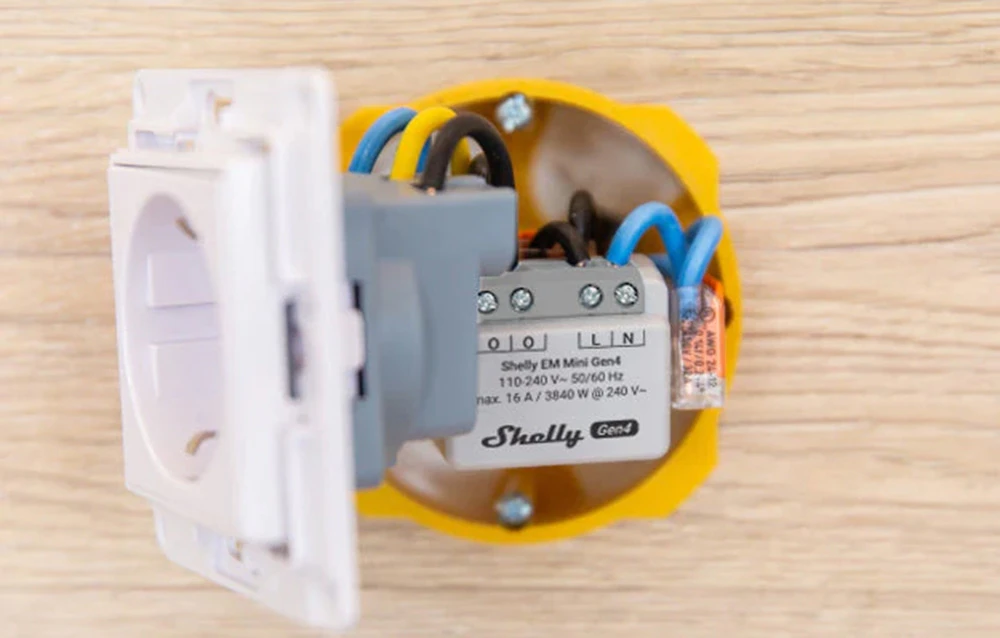 Un contor de energie alb montat într-o cutie electrică galbenă din perete, conectat la o priză cu cabluri.